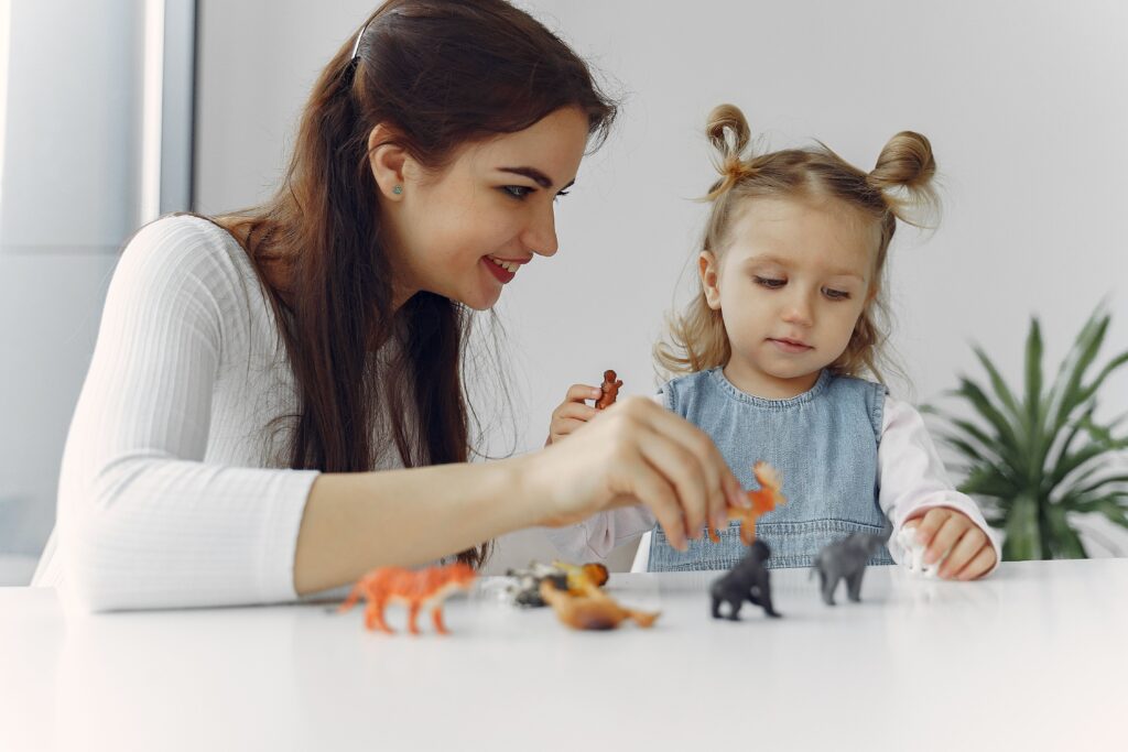 Babysitting & Childminding Adorable little girl sitting at white table with mother while playing with small toy wild animals for early education and development during spending time together at home