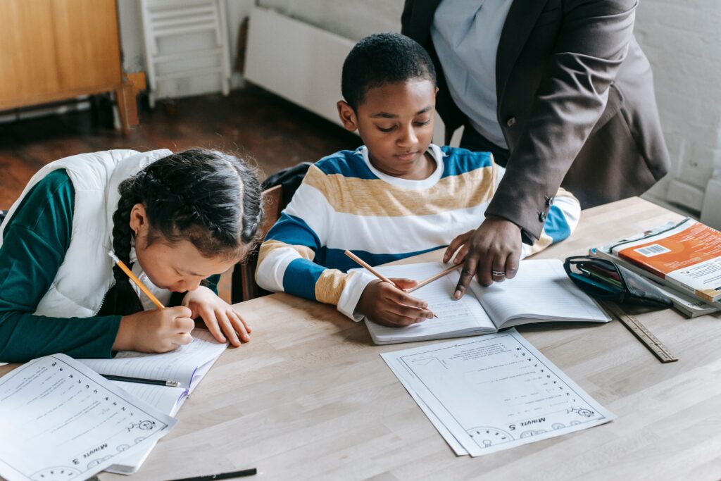 Homework Help High angle of crop unrecognizable black female teacher explaining task to focused little schoolboy sitting at desk near attentive Asian classmate writing in notebook